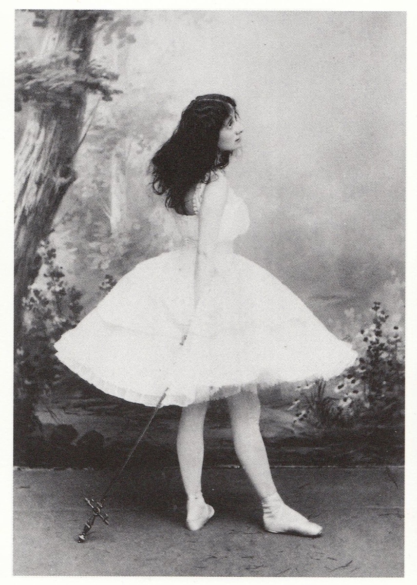 Anna Pavlova en Giselle, de profil dans la scène de la folie : tutu blanc, cheveux dénoués, l'épée abaissée vers le sol devant un décor de forêt peint.