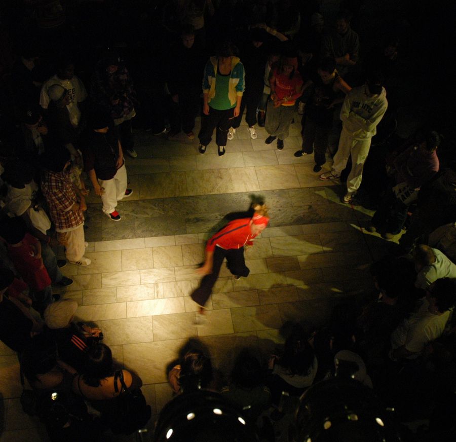 Vue plongeante sur un cercle de personnes debout, serrées les unes contre les autres dans la pénombre. Au milieu, une zone de sol éclairée reste vide, sauf une danseuse seule, en mouvement, penchée en avant.