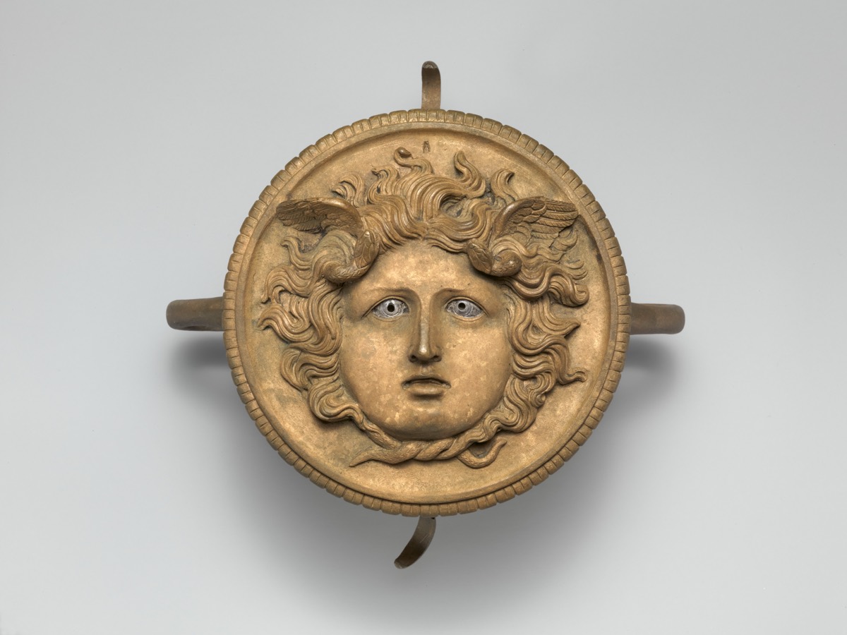 Ornement de timon de char romain : un visage de Méduse en bronze, calme et frontal, des ailes dans la chevelure, les yeux incrustés d'argent, deux serpents noués sous le menton.