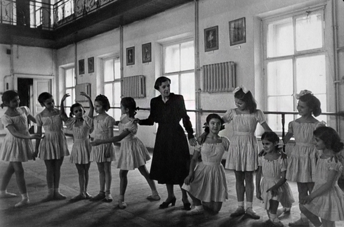 Une maîtresse de ballet en robe sombre, au centre d'une classe de jeunes élèves en tuniques claires, dans un grand studio à hautes fenêtres bordé d'une barre.
